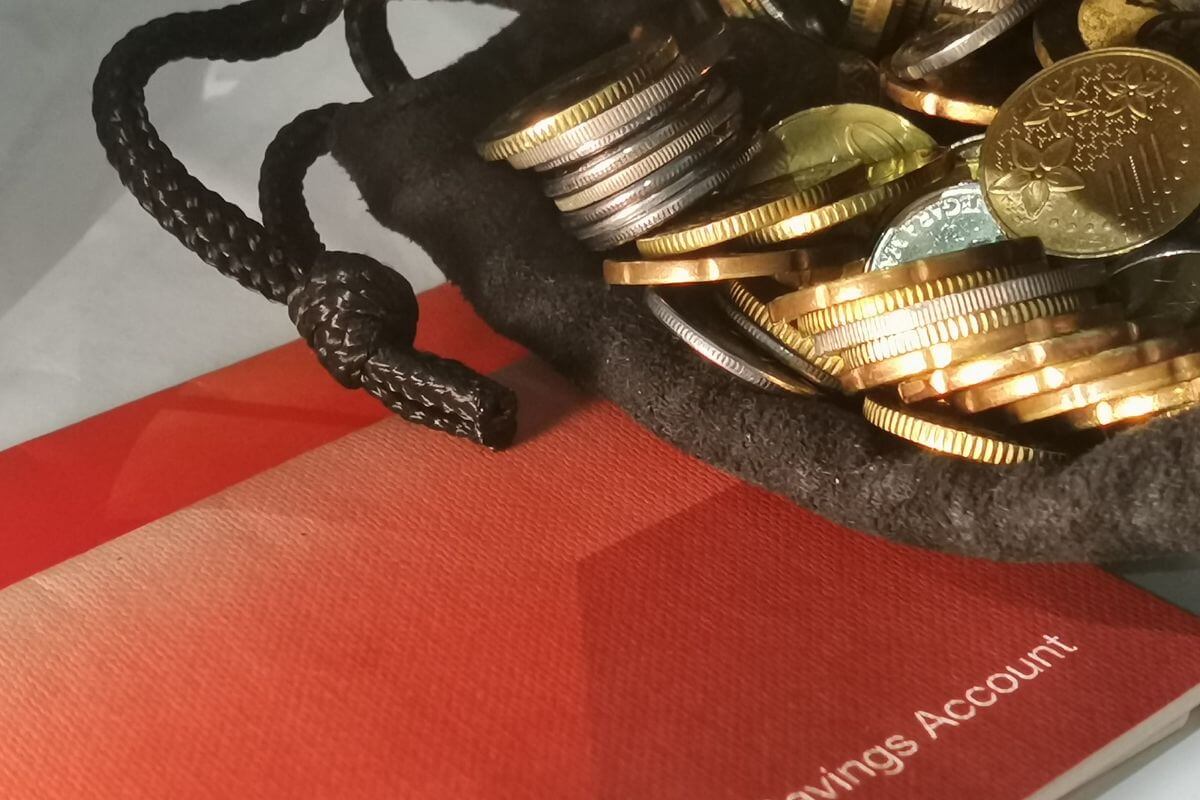 The picture shows a black pouch with coins and a bank book referring to opening a separate savings account for emergencies and future goals that give you a safety net while building good habits.