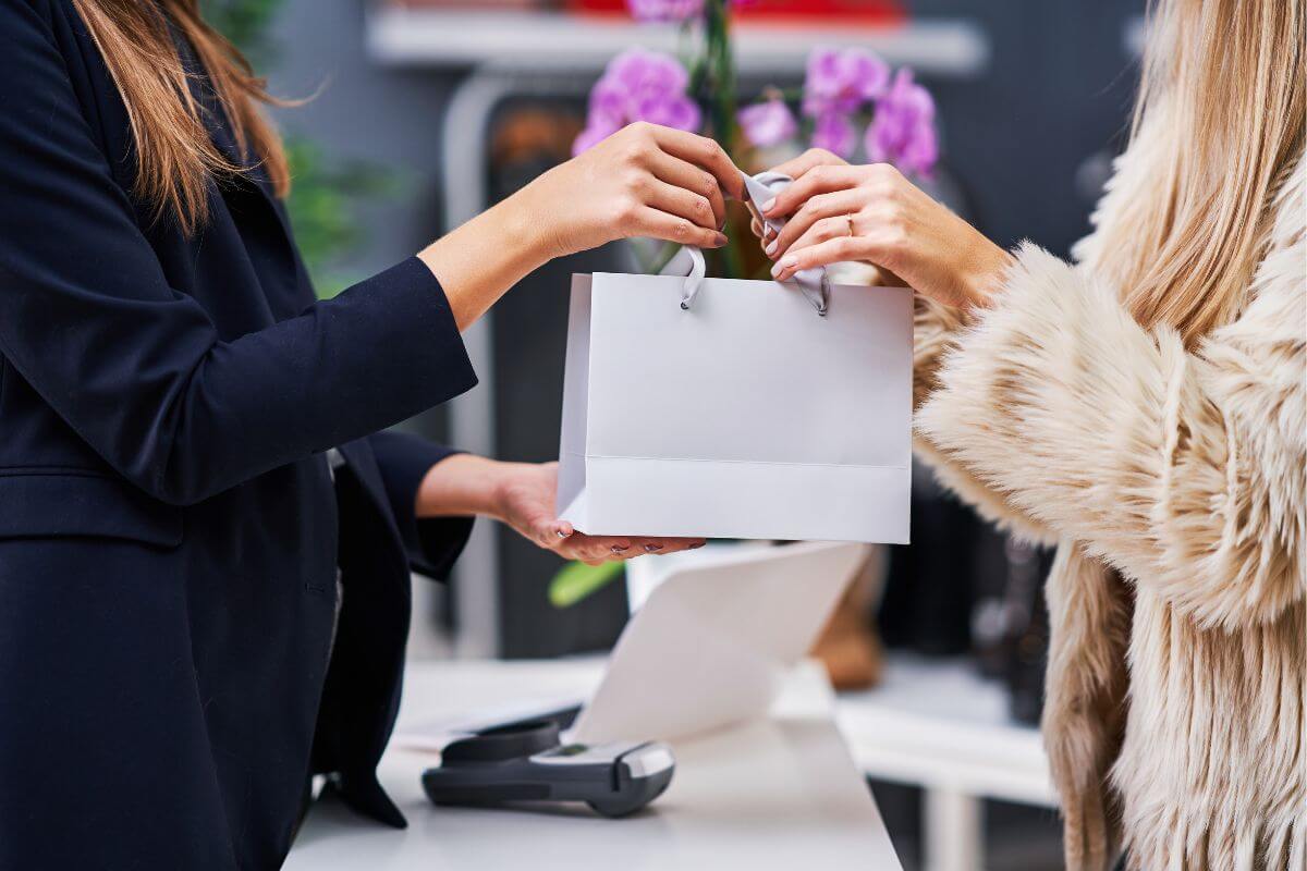 The image shows a cashier handing a shopping bag to a customer indicating to limit impulse buying by following the 24-hour rule helps you avoid overspending on things you don't need.