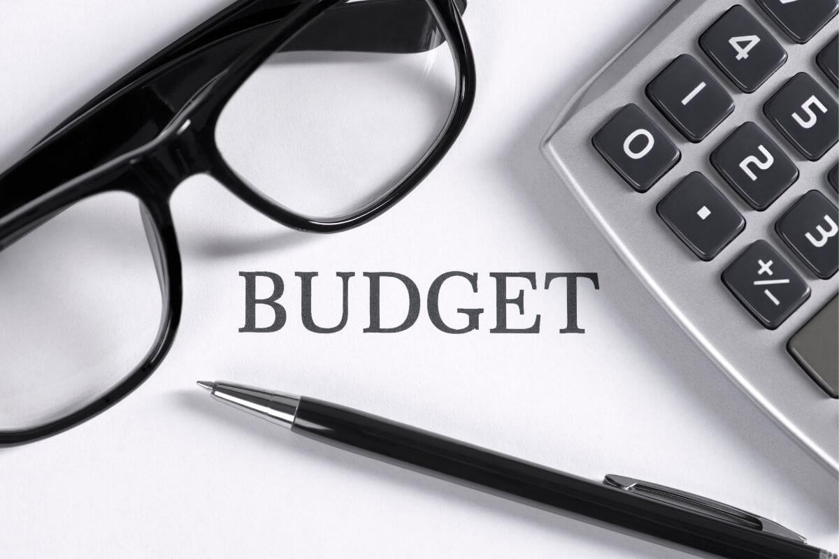 The photo shows a calculator, a pen, an eyeglass, and a piece of paper with a note indicating to continuously reassess your budget and adjust as needed to keep up with changing expenses and goals.