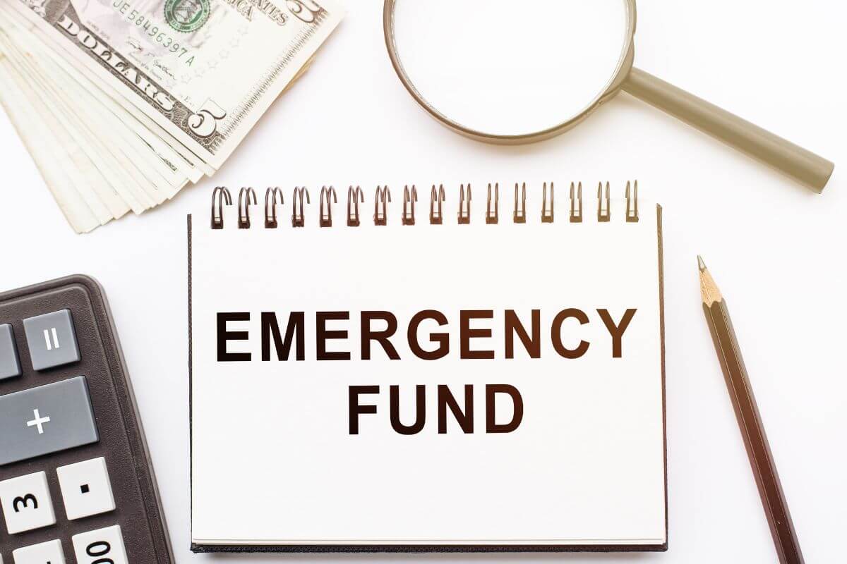 The photo tells about dollar bills, a magnifying glass, a pen, a calculator, and a a notebook with notes indicating to no emergency fund may turn to debt and financial stress.
