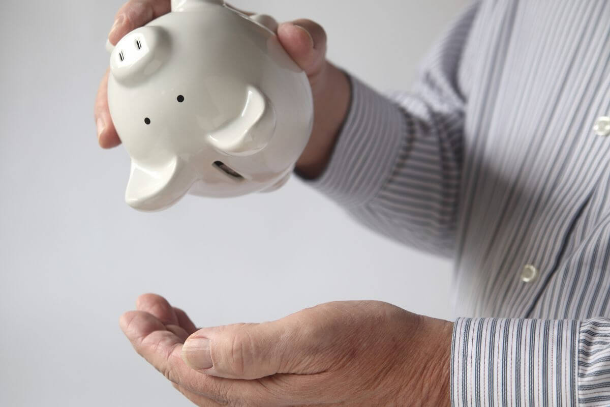 The image shows an old man shaking a piggy bank upside down without single coins referring to a no savings leaves you vulnerable.