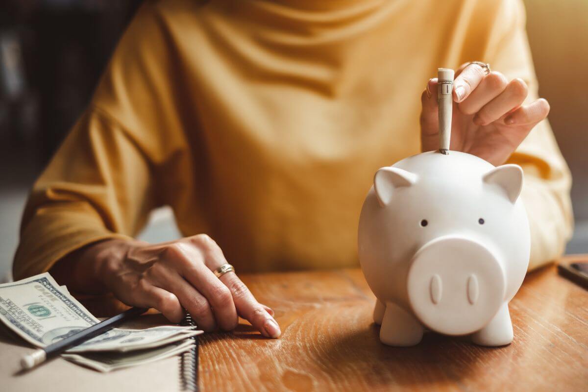 The image shows a notebook, a pen, dollar bills, and a girl dropping a dollar bill on a piggy bank indicating to use the reverse budgeting method to manage the rest of your spending guilt-free.