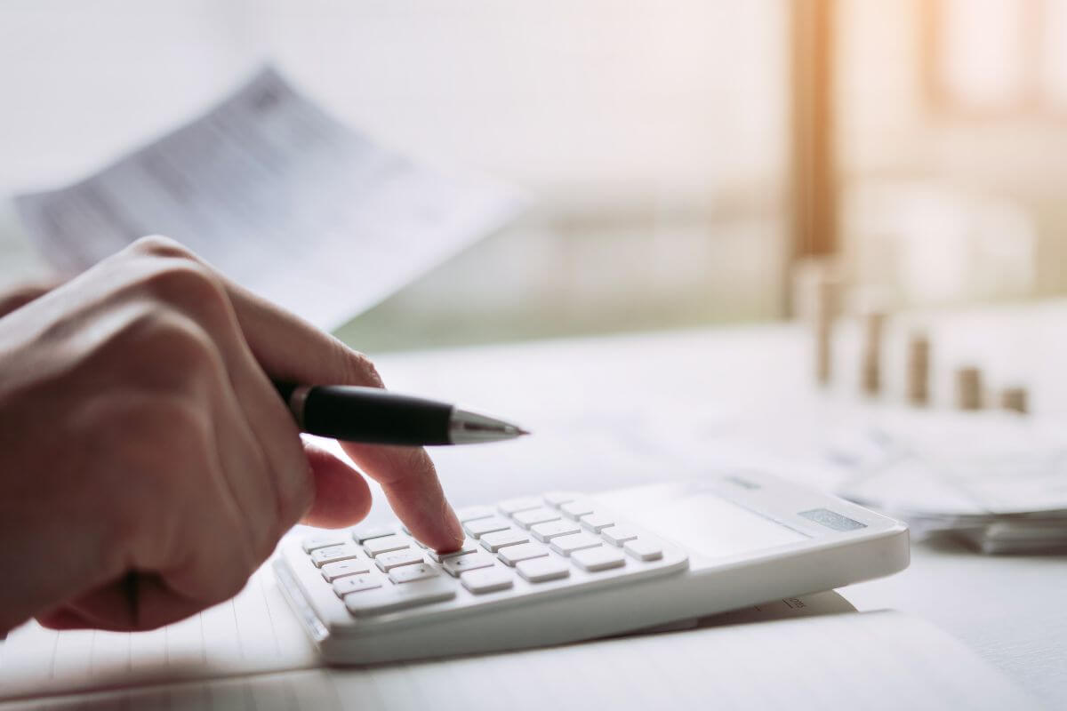 The photo tells about coin stocks, dollar bills on the table, and a man's hand calculating while holding a paper referring to learning more ideas to budget better today to take control of your money, save more, and work toward financial independence.