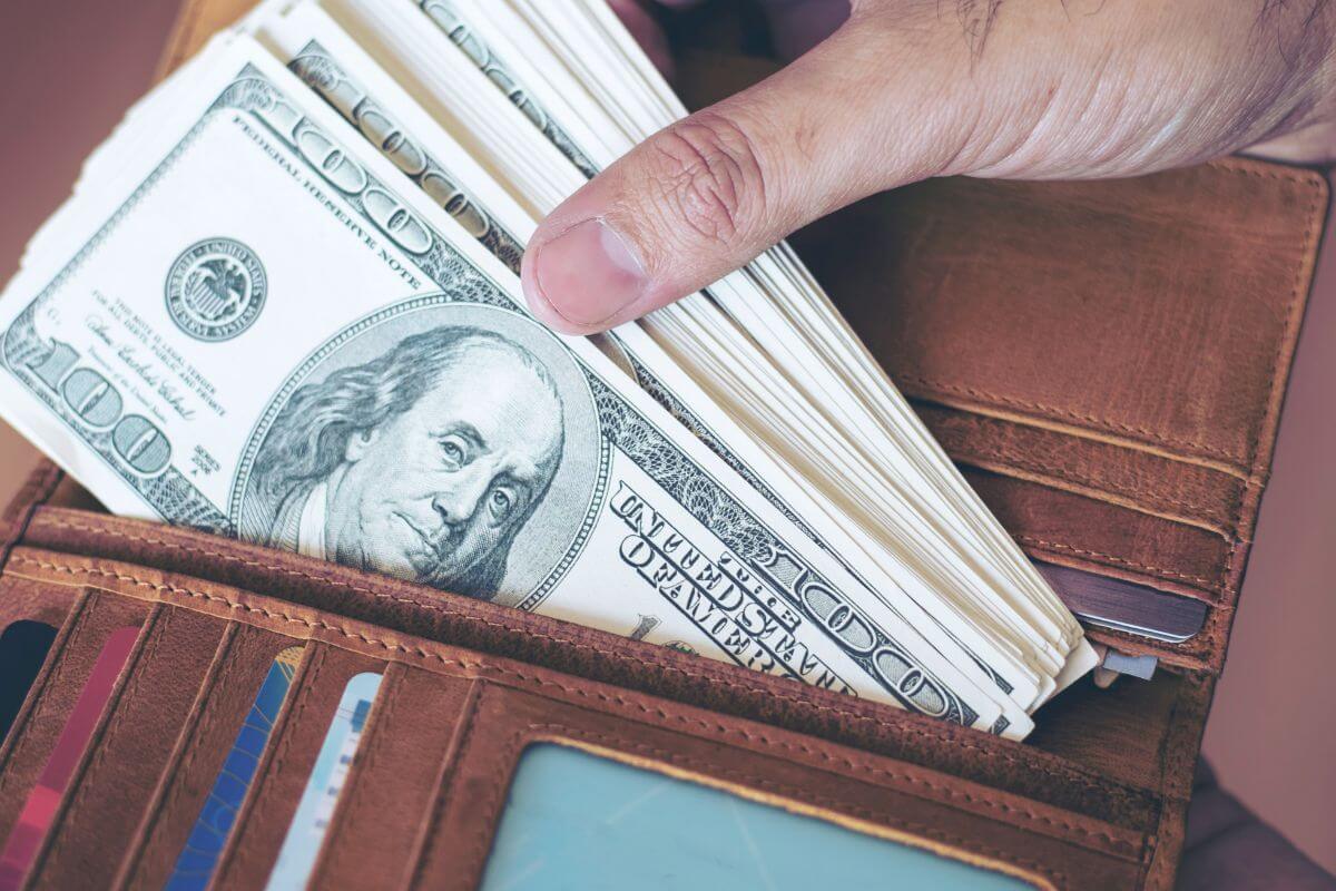 The image of a wallet with cards and a man's hand pulling out dollar bills refers to committing to no spend challenge months regularly and becoming more mindful of where your cash goes.