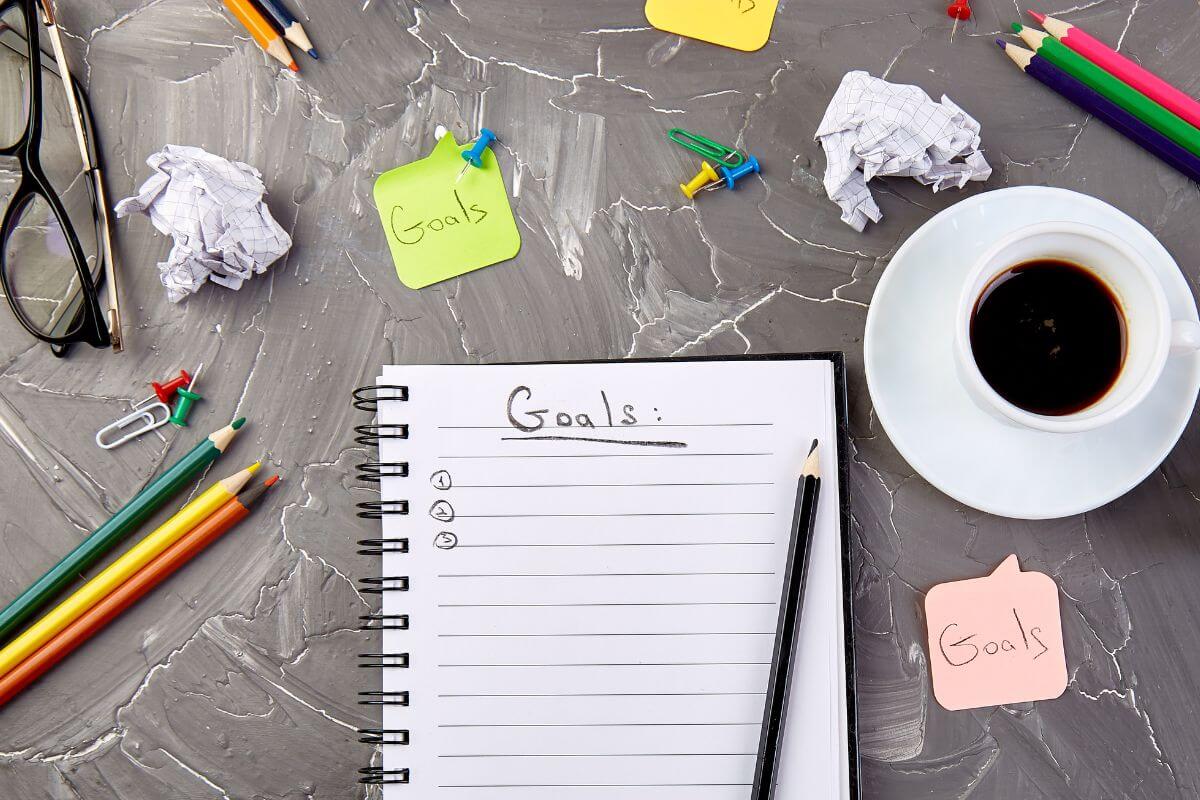 The image shows crumpled papers, an eyeglass, pens, a coffee, pins,  small pieces of paper with notes, and a notebook with notes indicating that setting short-term goals keeps you motivated to keep pushing forward.