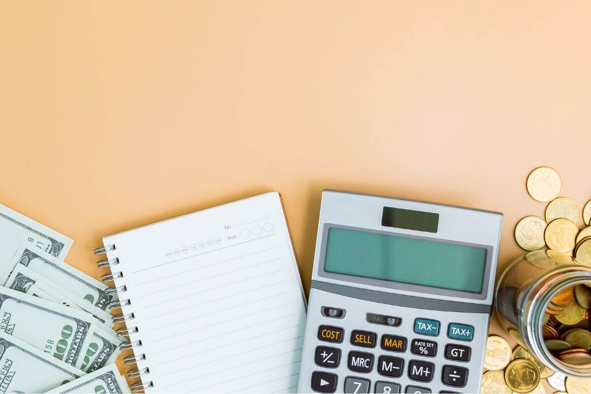 The photo of dollar bills, a notebook, a calculator, coins on the table, and a jar filled with coins referring to setting long-term goals helps you focus on what matters most.