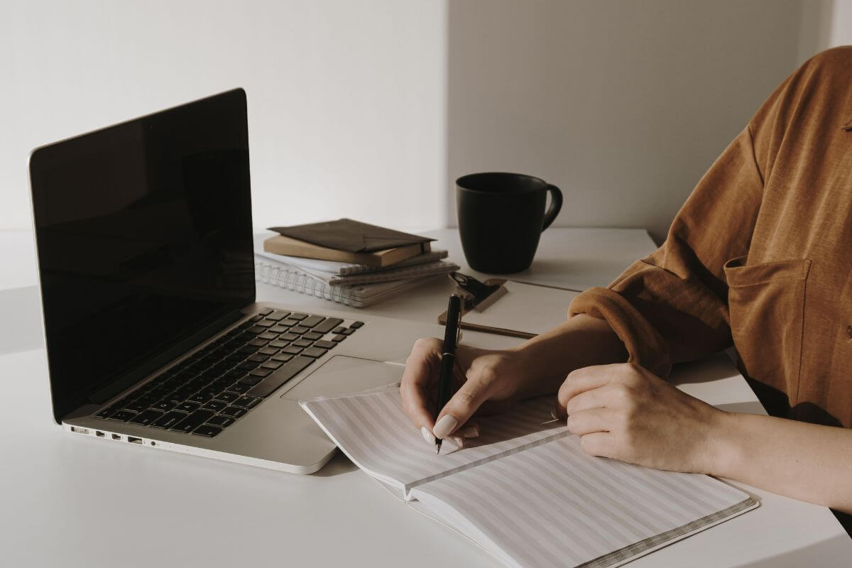 The photo of a cup, notebooks, a clipboard, a laptop, and a girl writing on the notebook means to set clear financial goals that help you stay focused and motivated.