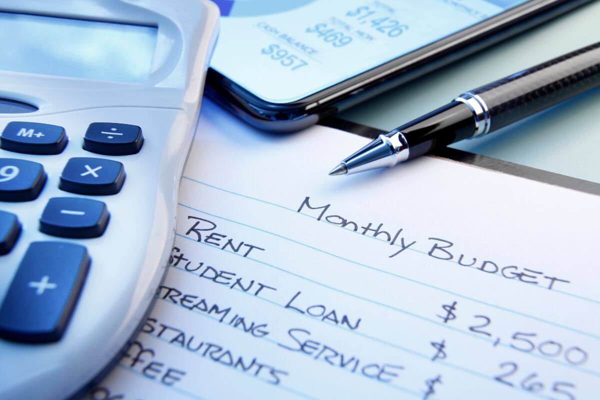 The picture shows a phone with a screen, a pen, a calculator, and a notebook with notes referring to prioritizing your monthly budget with investing first which keeps you consistent and builds your portfolio faster.