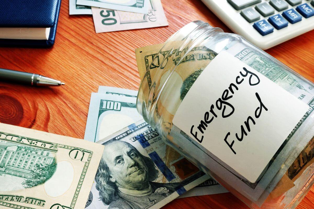 The picture tells about dollar bills on the table, a pen, a notebook, a calculator, and a jar with dollar bills and notes referring to building an emergency fund before you invest prevents you from pulling money out of investment.