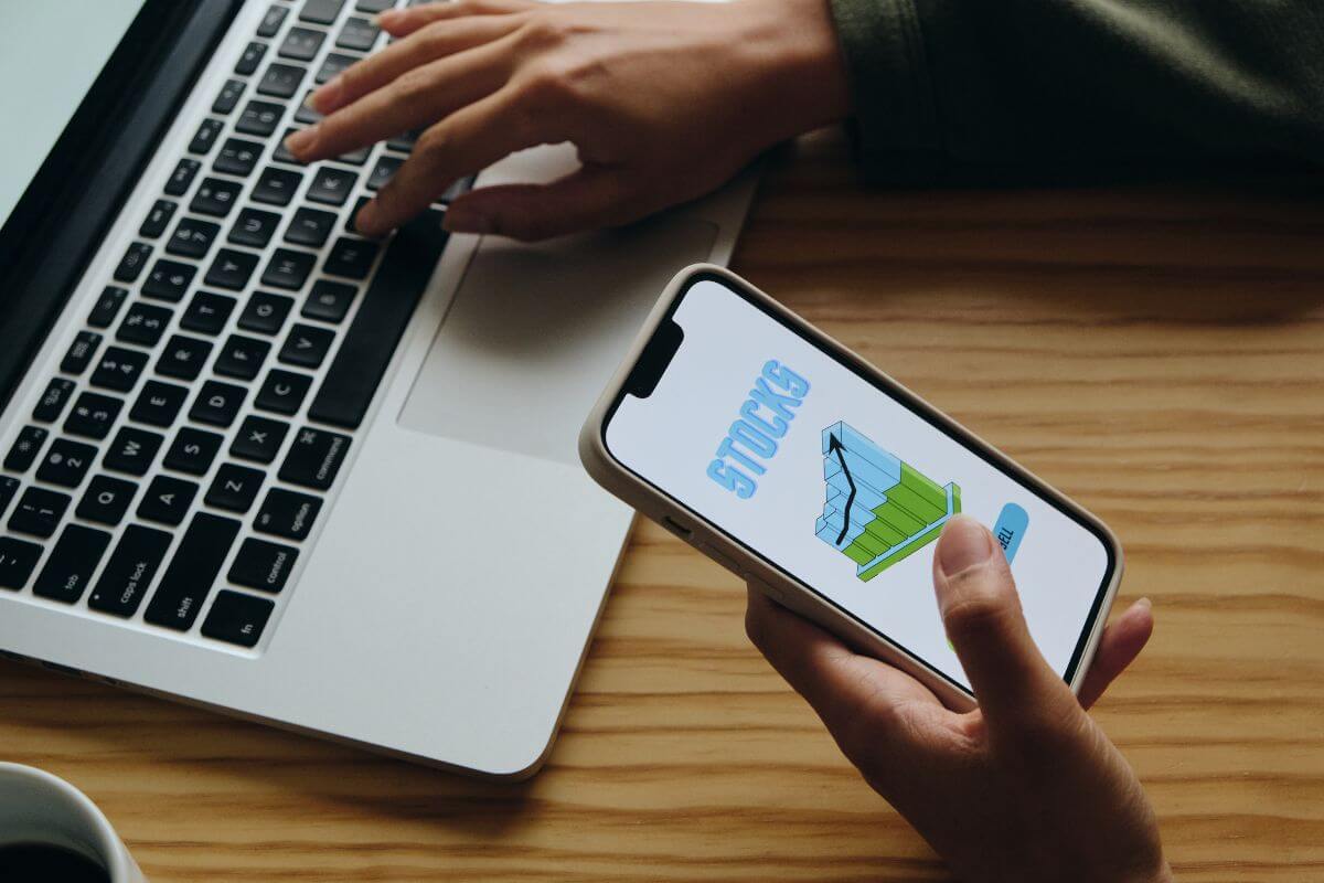 The image shows a girl's hand using a phone with a screen display while using also a laptop which means learning basic investing terms and growing your knowledge over time.