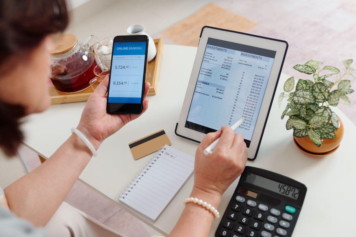 The picture shows a plant, tea, a credit card, a calculator, and a girl using a phone with a screen display while checking her tablet device indicating to open a brokerage account to start building your investments.