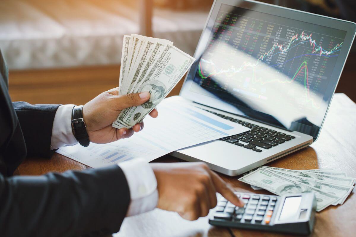 The image tells about a piece of paper with graphs, dollar bills on the table, a laptop with a screen display, and a man's hand holding dollar bills while calculating indicating to diversify your investments to reduce risk to protect yourself if one performs poorly.