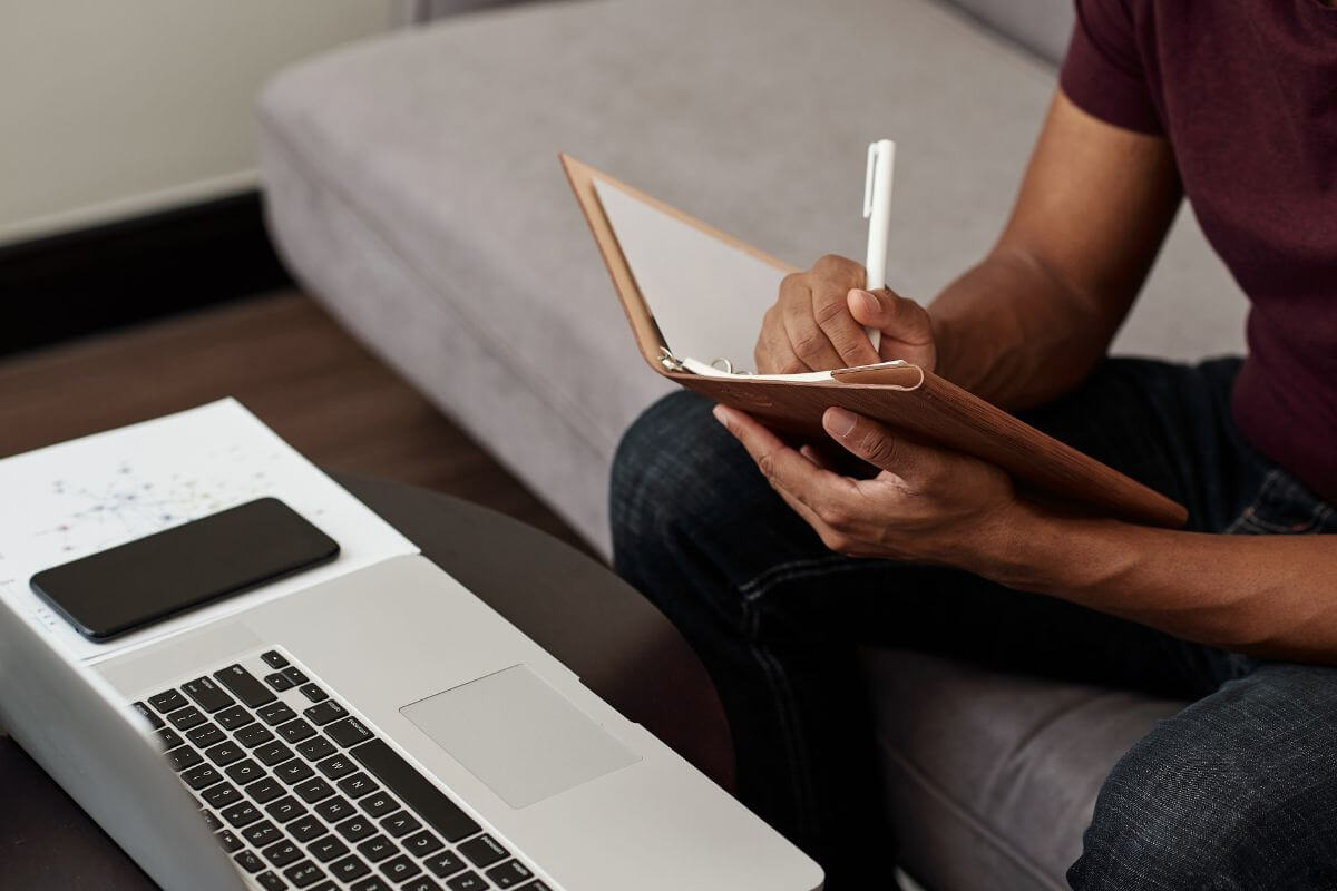 The picture tells about a phone, a laptop, and a man jotting down on a notebook while watching online to continue learning about investing to be more confident in making smart investment choices.