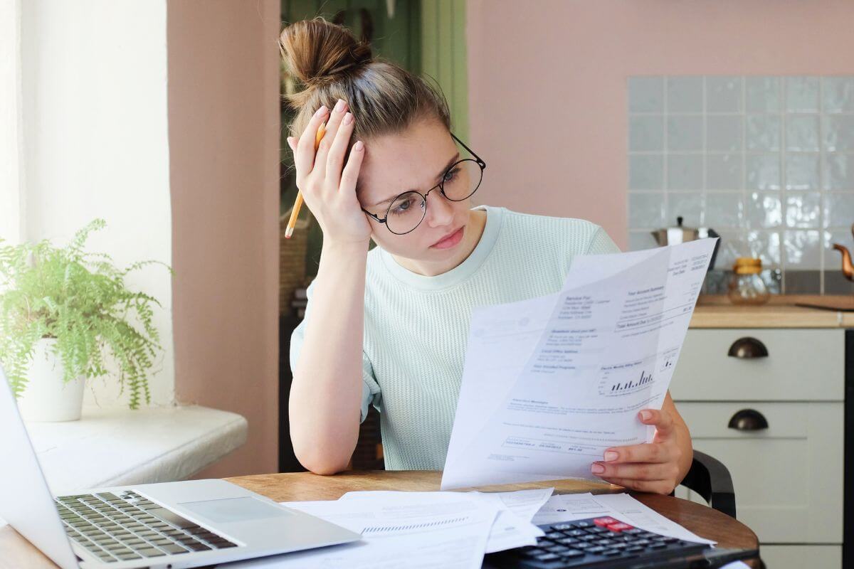 The photo tells about papers on the table, a calculator, a laptop, and a girl looking at a paper while holding her head which means to pay down high-interest debt to free up money for savings and building wealth over time.