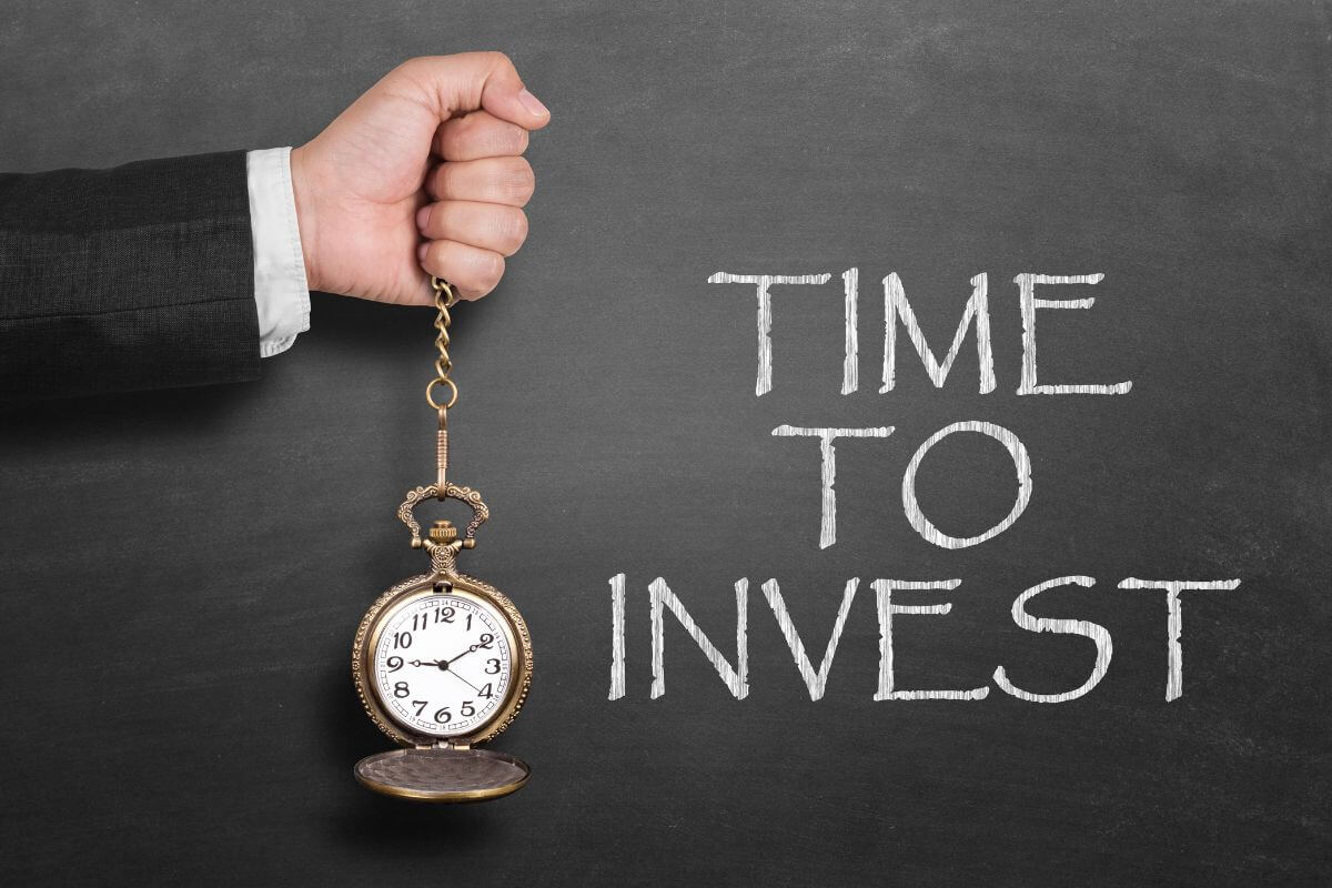 The photo shows a man's hand holding a clock and a blackboard with chalk notes indicating to don't be too afraid to invest that you never start and become financially stable.
