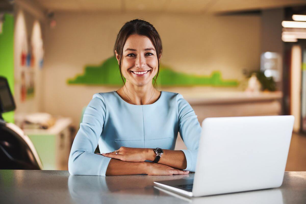 The image shows a laptop on the table and a girl smiling which refers to become a virtual assistant to earn money from the comfort of your home.