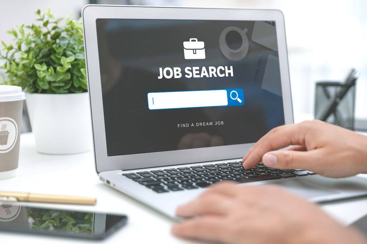 The photo tells about a plant, a coffee, a phone, and a man's hand typing on a laptop with a screen display to find a high-paying job to help you save more and grow your net worth.