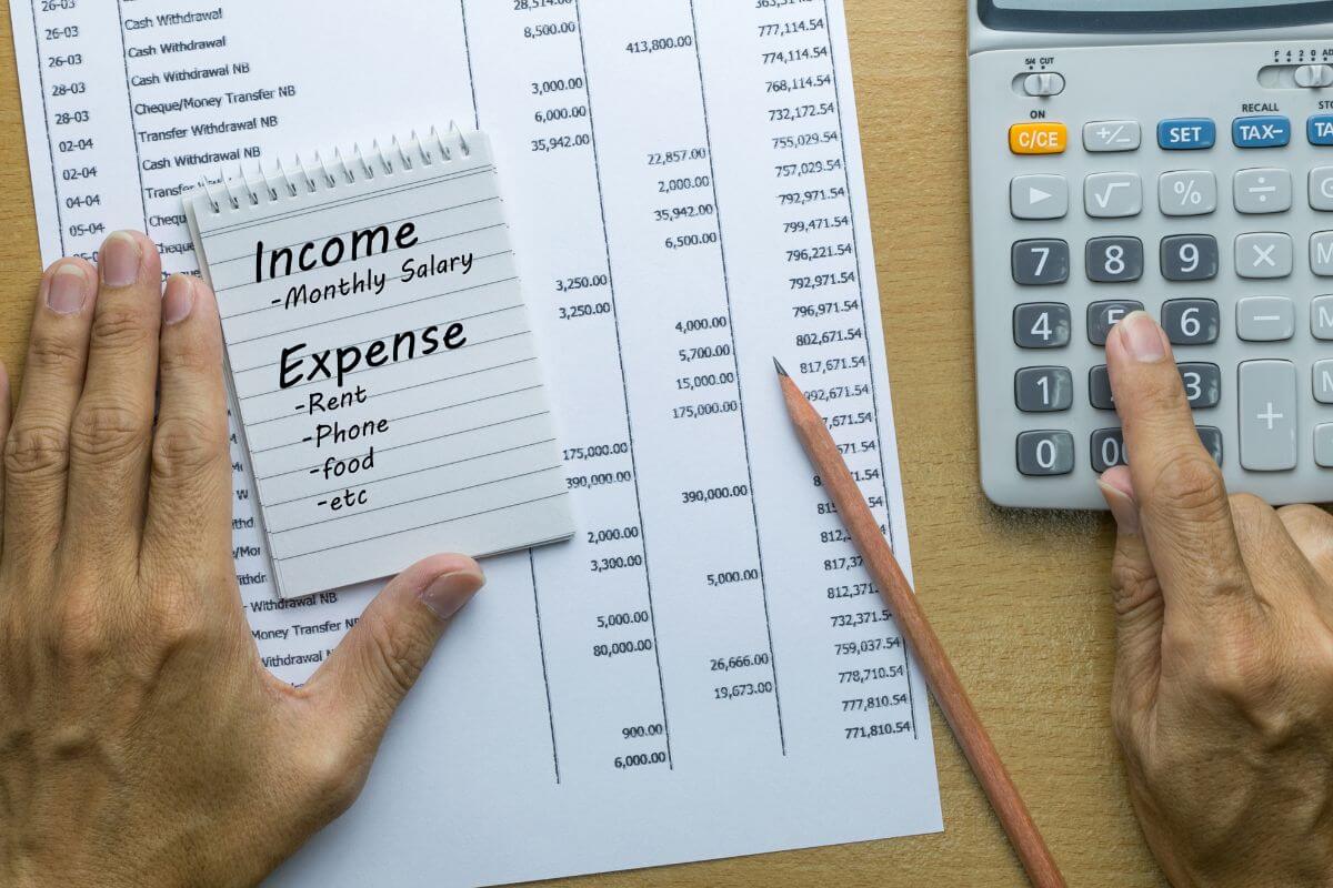 The picture tells about a piece of paper with notes and numbers, a pen, a mini notebook with notes, and a man's hand calculating which means to plan for tracking income and expenses that help keep your spending in check and ensure you can save for what really matters.