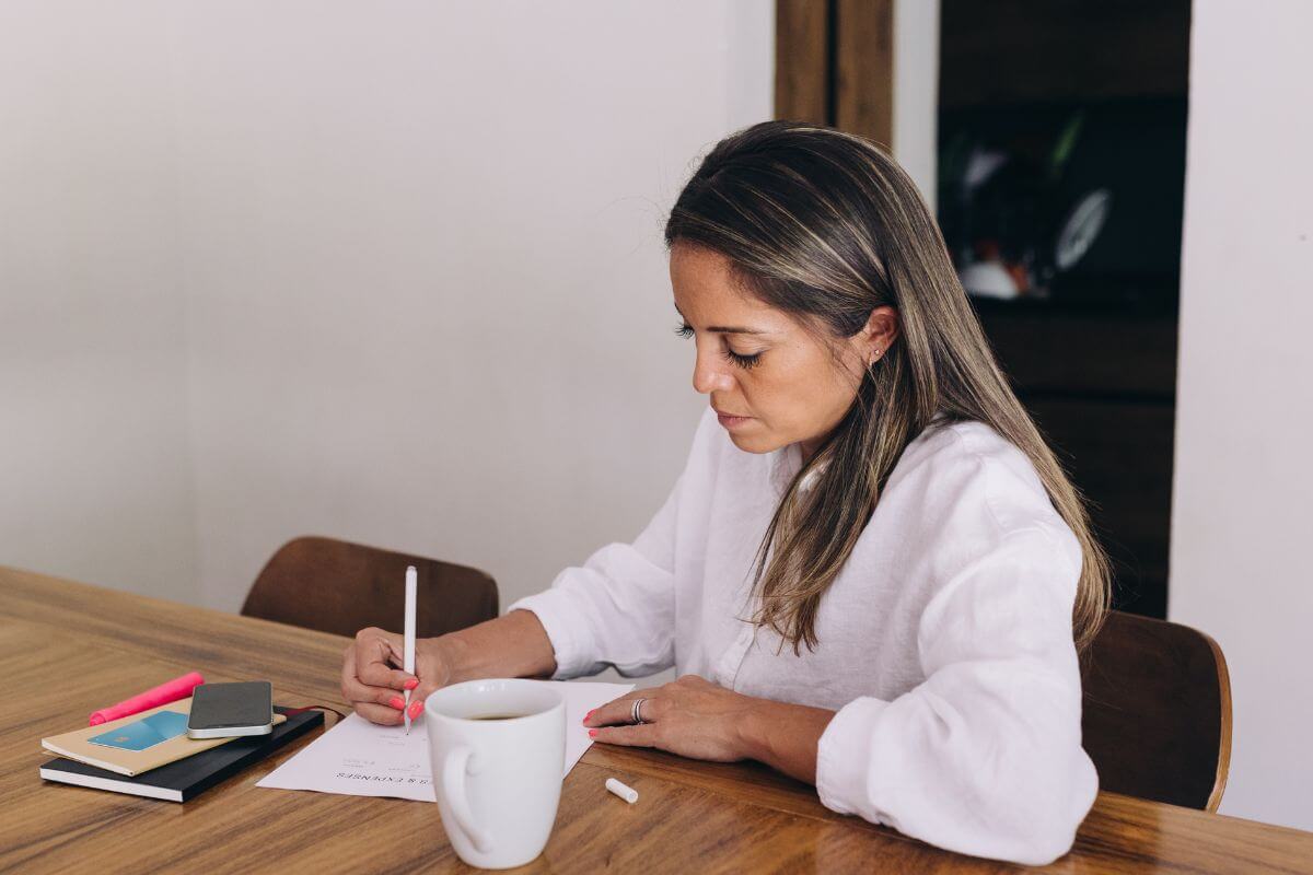 The photo shows a phone, notebooks, a coffee, and a girl writing on the paper which refers to tracking monthly expenses to give you a clear picture of your spending.