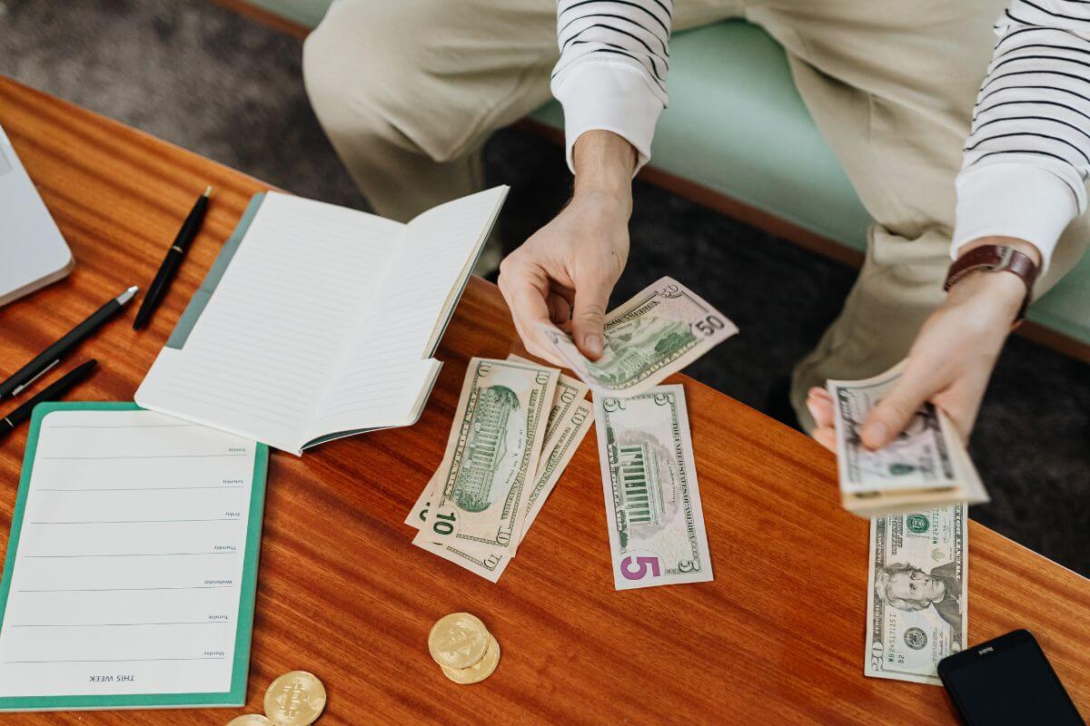 The image of pens on the table, notebooks, coins, a phone, and a man's hand grouping the money means to categorize spending to see where the money goes and what can be adjusted.