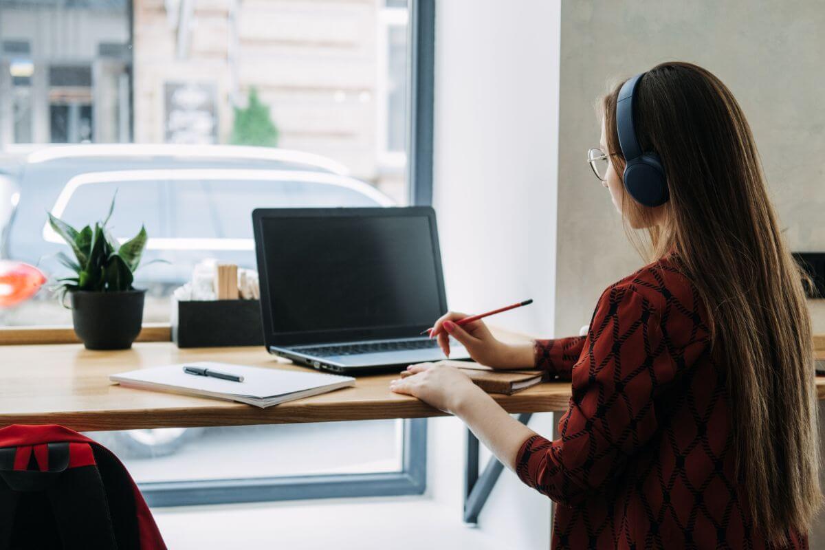 The photo of a plant, a notebook, a pen, a laptop, and a girl sitting while using a laptop to jot down refers to taking on a side hustle and growing savings for financial independence.