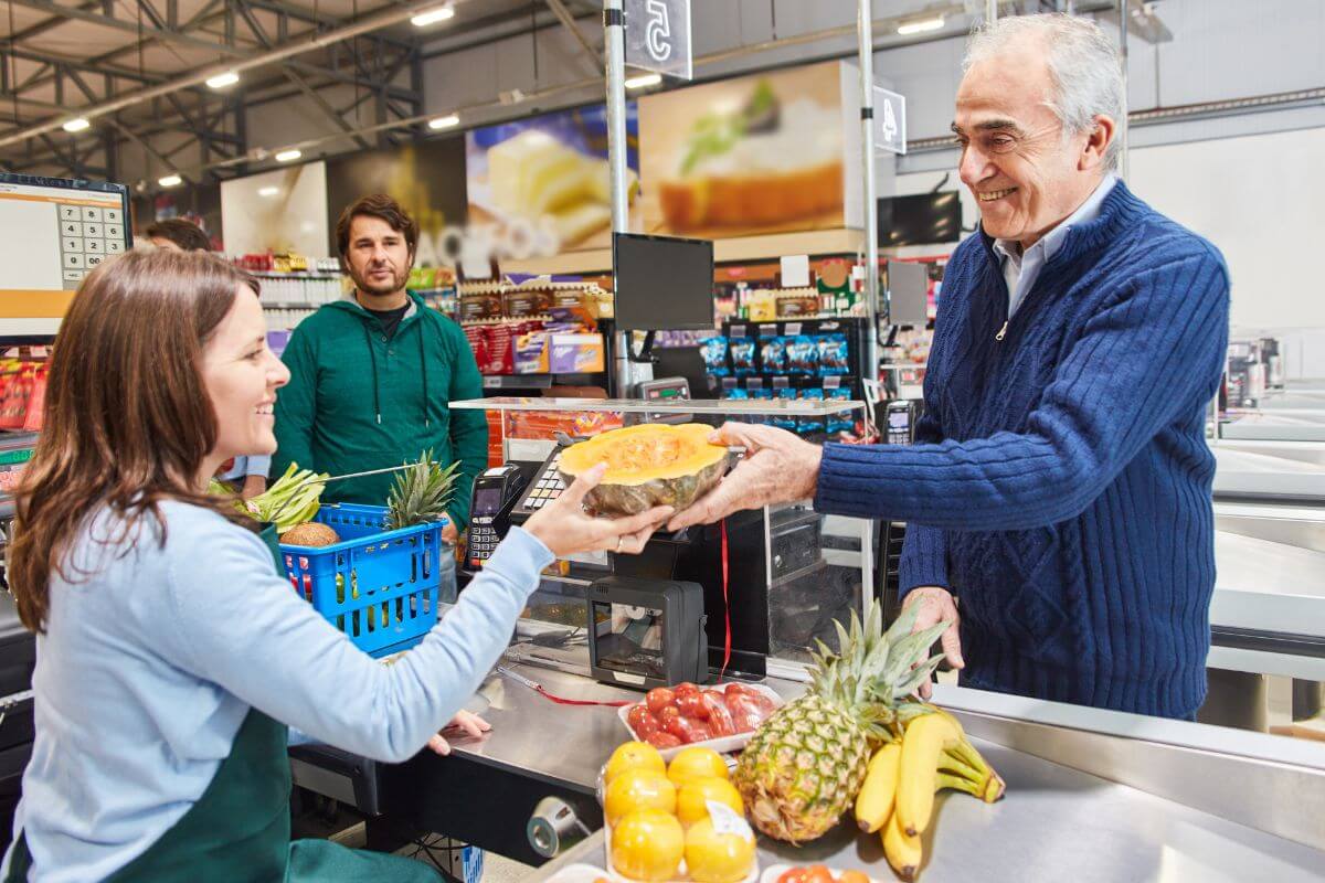 The image shows a senior buying fruits and vegetables at the store and handing the squash to her while the other customer is waiting in the next line referring to shop during senior discount days to save money on your weekly food budget.