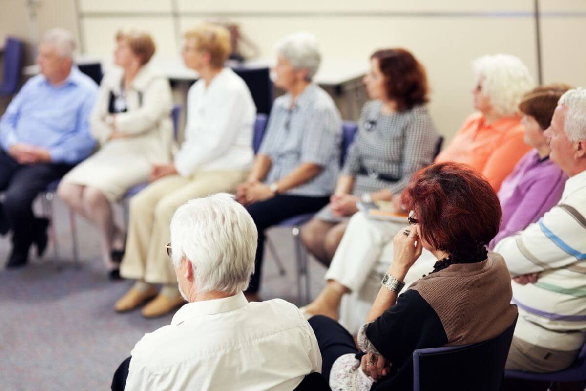 The picture shows seniors at the venue sitting while listening, indicating that they should sign up for free or discounted classes at community centers to learn and stay active without high costs.