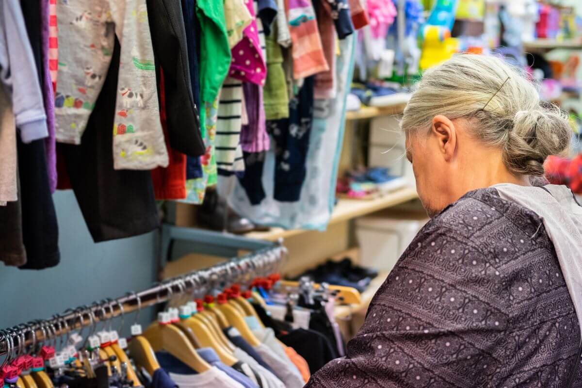 The picture shows a senior picking clothes at the thrift store indicating to shop secondhand for clothing to save money on essentials and extras. 