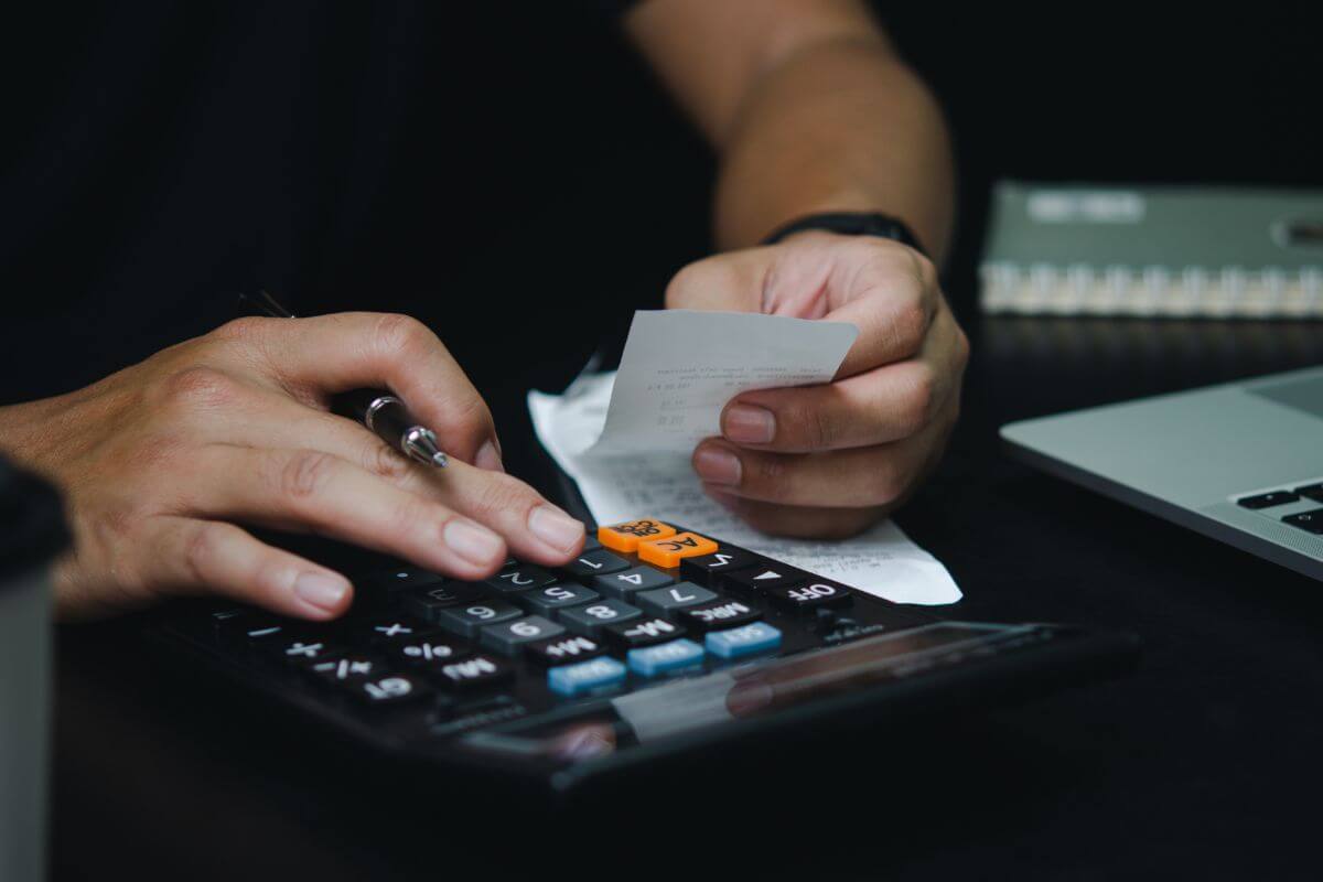 The image of a laptop, a notebook, and a man's hand calculating a receipt which means to identify overspending in non-essential categories and cutting back it leaves you with extra cash to pay down debt or add to savings.