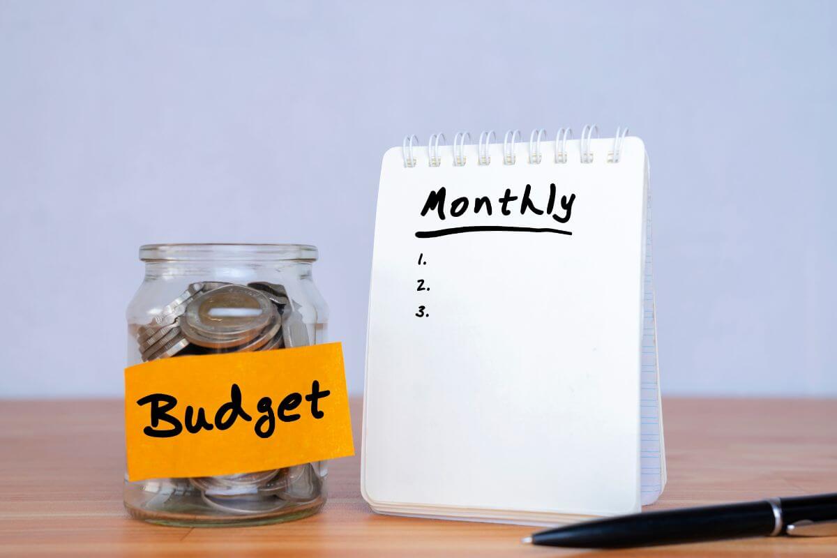 The image tells about a pen, a notebook with notes, and an ajar filled with coins and a note referring to creating a realistic monthly budget helps you save or pay off debt without feeling overwhelmed.