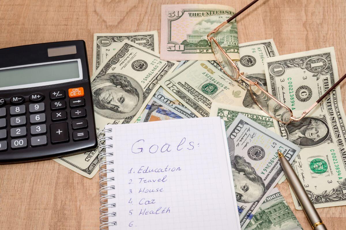 The picture shows a calculator, a pen, an eyeglass, dollar bills, and a notebook with notes indicating to set financial goals to spend less and put more toward what really matters.