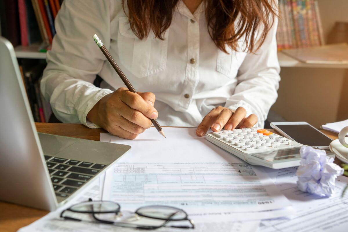 The photo shows a laptop, an eyeglass, papers, a phone, a calculator, and a girl writing on the papers which means tracking progress regularly to see how far you've come and help you build lasting habits.