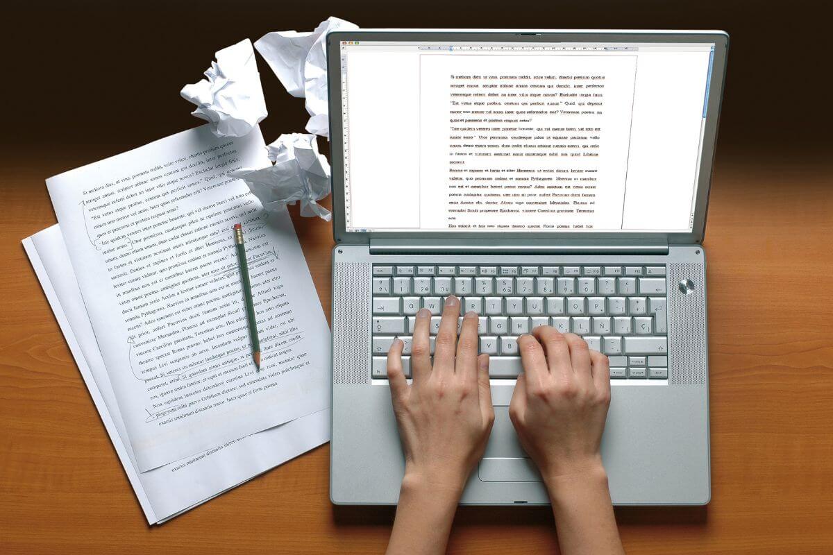 The image shows papers on the table, a pen, crumpled papers, and a girl's hand typing on a laptop which means to start freelance writing to have a steady and lucrative income sourcce.
