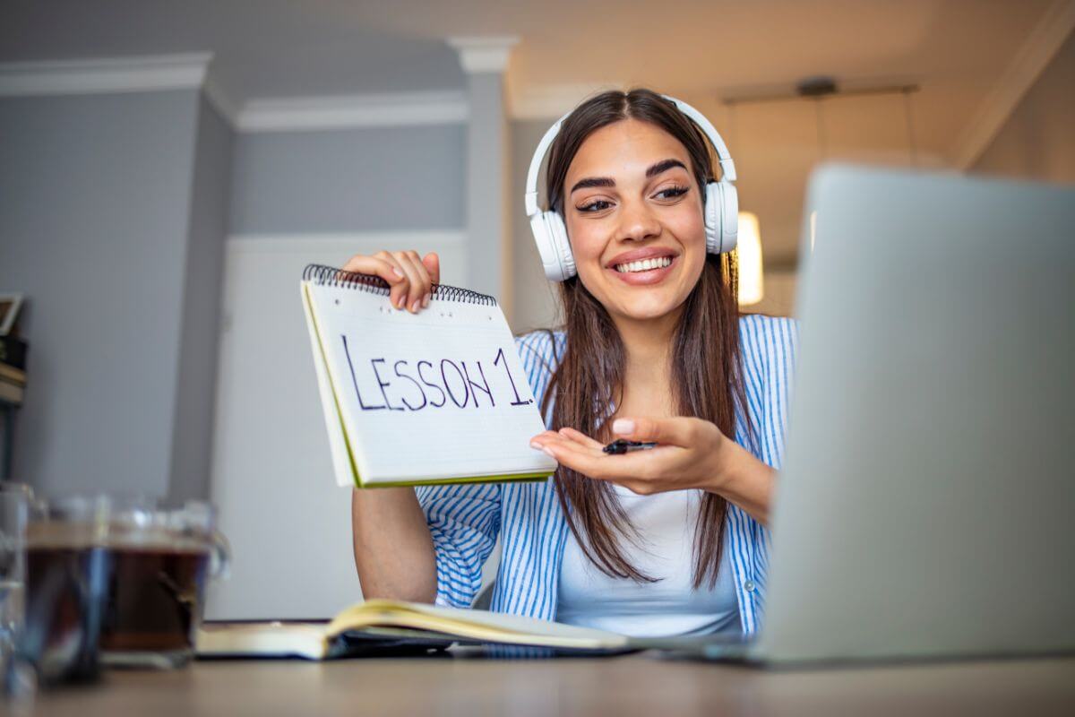 The photo shows a book on the table and a girl wearing a headset while holding a notebook with notes showing on the laptop referring to provide online tutoring is a rewarding way to earn from home while helping others learn.