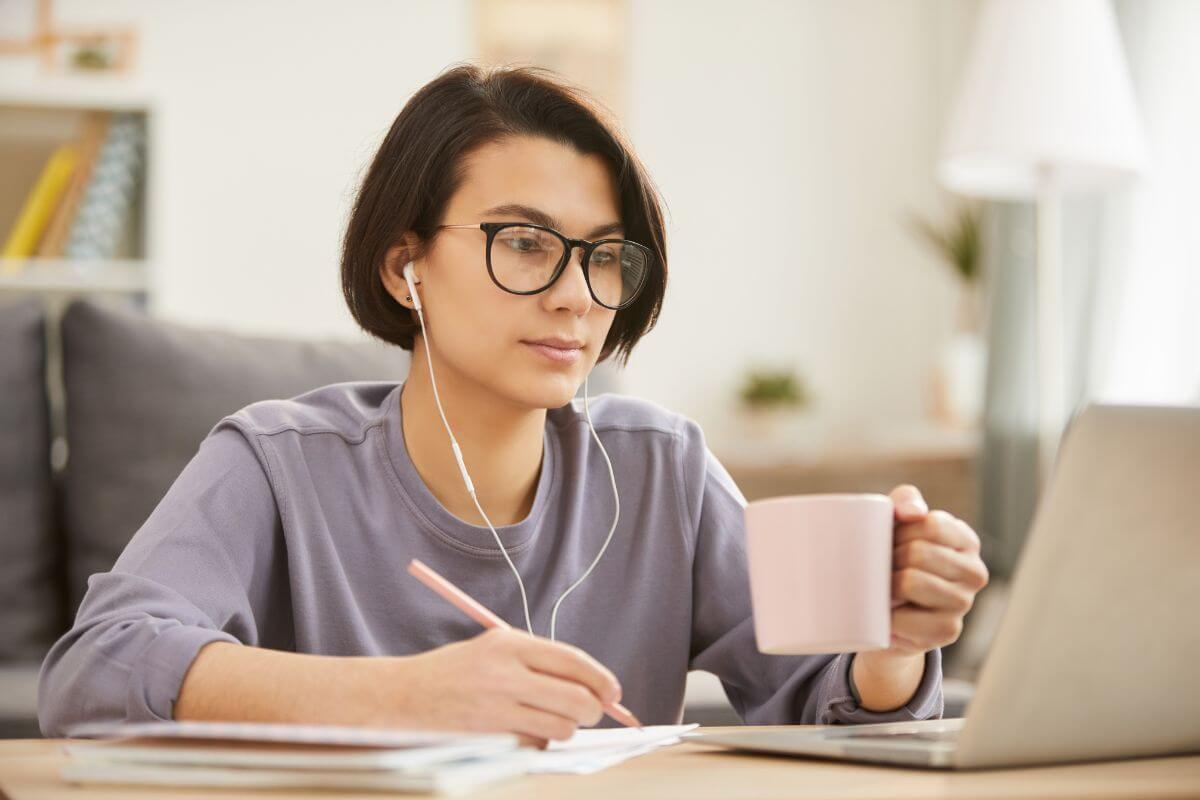 The picture tells about papers in a notebook and a girl writing on the paper and looking at the laptop while having a coffee at home indicating to work as a transcriptionist which is an easy way to earn a steady income with minimal investment. 