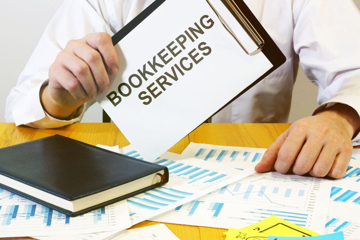The photo of papers with graphs, a notebook, and a man holding a clipboard with notes indicating to provide bookkeeping services which is a dependable way to earn a professional income from home.