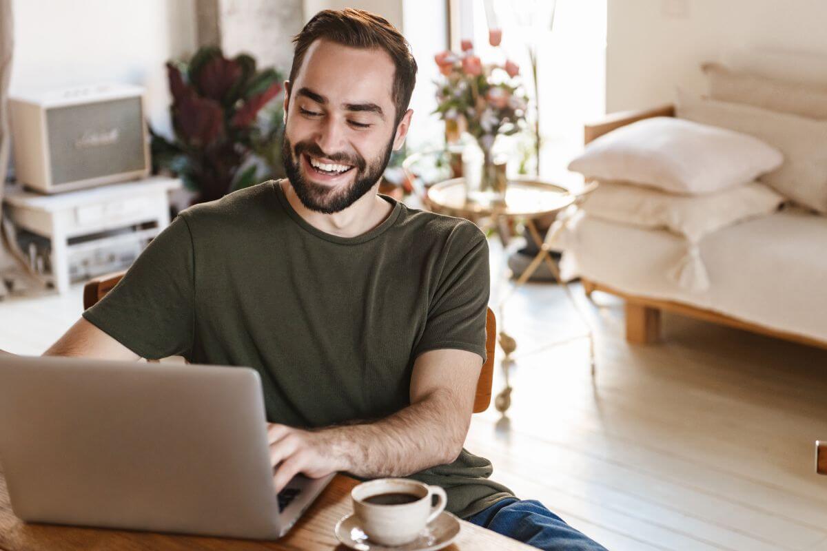The picture shows a couch with pillows, flowers on the table, a coffee on the table, and a man smiling while using a laptop referring to learning to make $2500 a month from home and turning ideas into reliable income streams.
