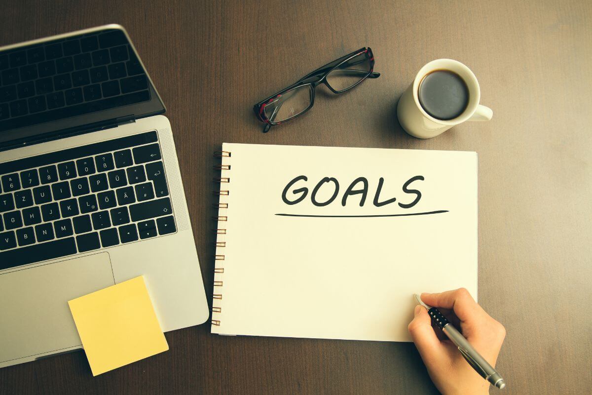 The picture of a laptop, an eyeglass, a coffee, a sticker note, and a girls hand writing on the notebook indicating to set your goal helps you stay motivated and the first step to financial success and guide to yoour savings.