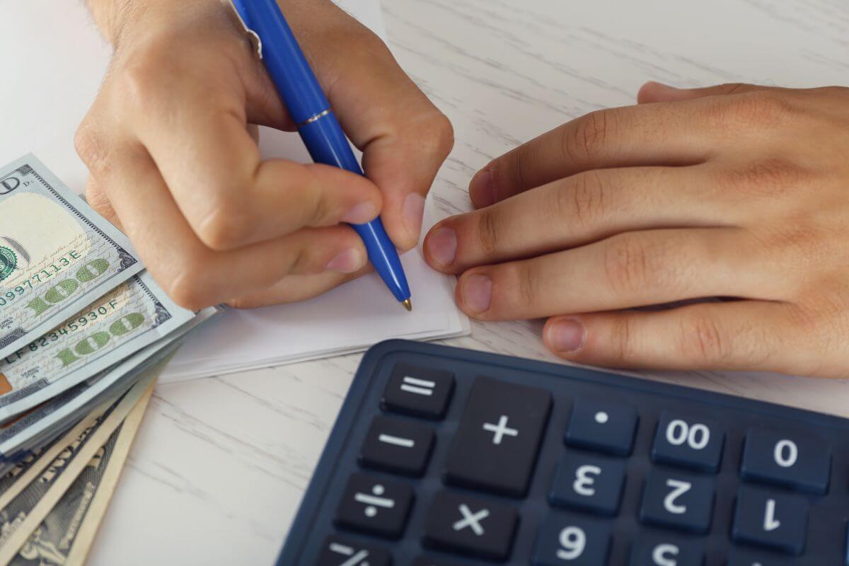 The image shows a calculator, dollar bills, and a man's hand and writing on the papers which means creating a budget to set money aside first helps you avoid overspending and ensures every dollar has a job.