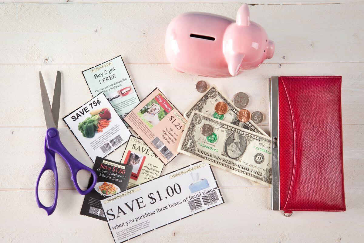 The photo shows a wallet, a piggy bank, a scissor, dollar bills, coins, and coupons on the table referring to become frugal that brings you closer to your $100k goal.