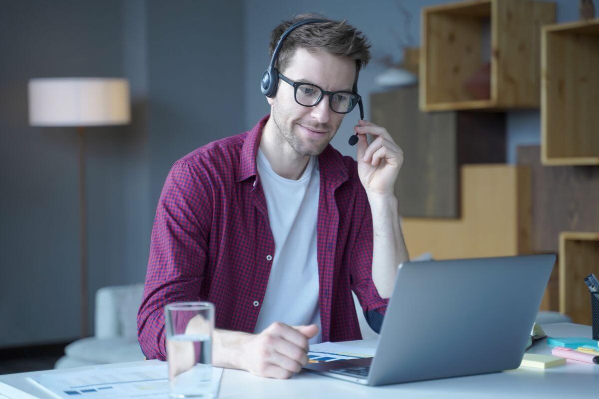 The image shows a glass of water, papers on the table, and a man using a headset with a microphone while looking at the laptop which means to offer consulting services that can turn into a high-paying and fulfilling job.