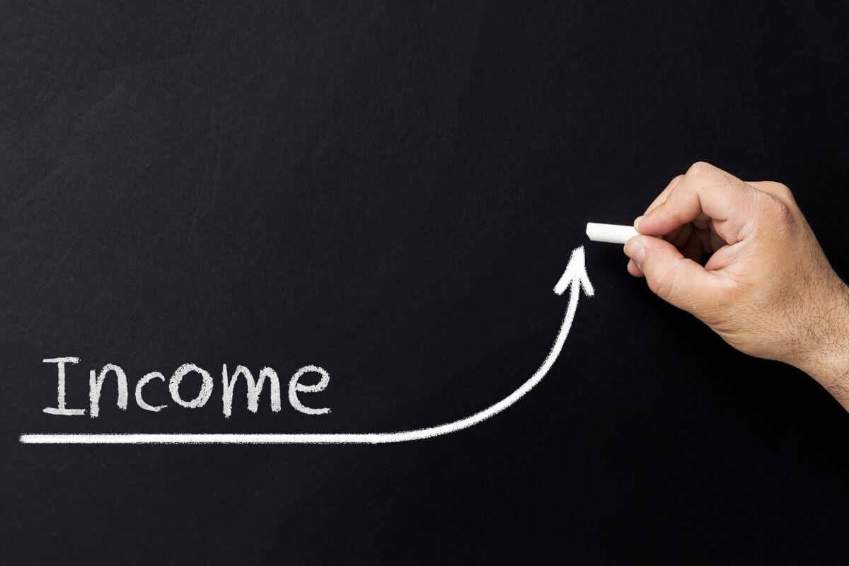 The picture of the man's hand holding a chalk writing on the blackboard of a rising arrow and a note referring to bringing in more cash flow. Increase your income 