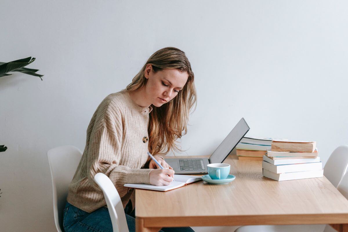 The picture of books on the table, a laptop, a coffee, and a girl writing on the notebook means to stay disciplined and staying consistent to make a difference.