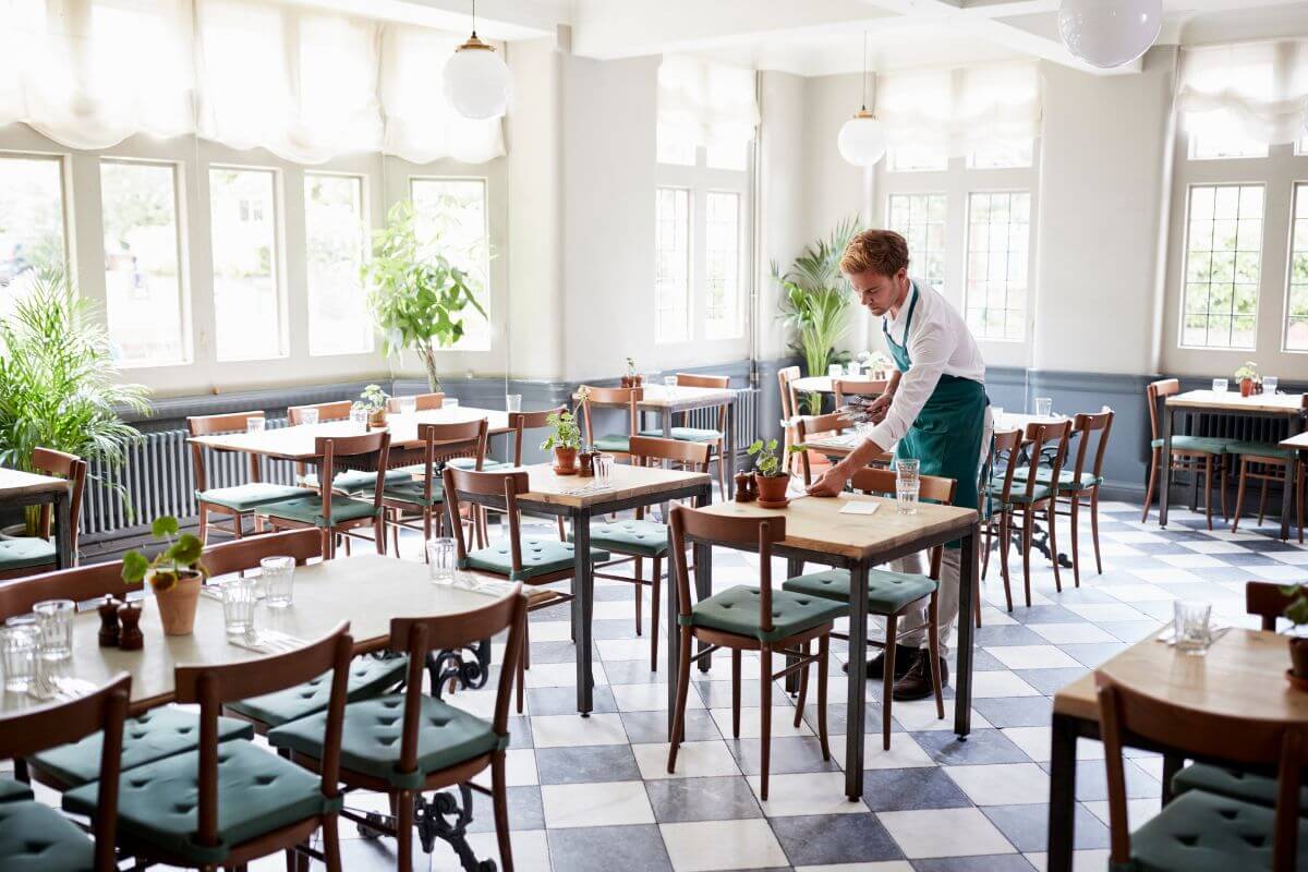 The image shows tables, chairs, plants, and a waiter cleaning the table indicating to become a mystery shopper for local businesses to get free products or meals along the way.