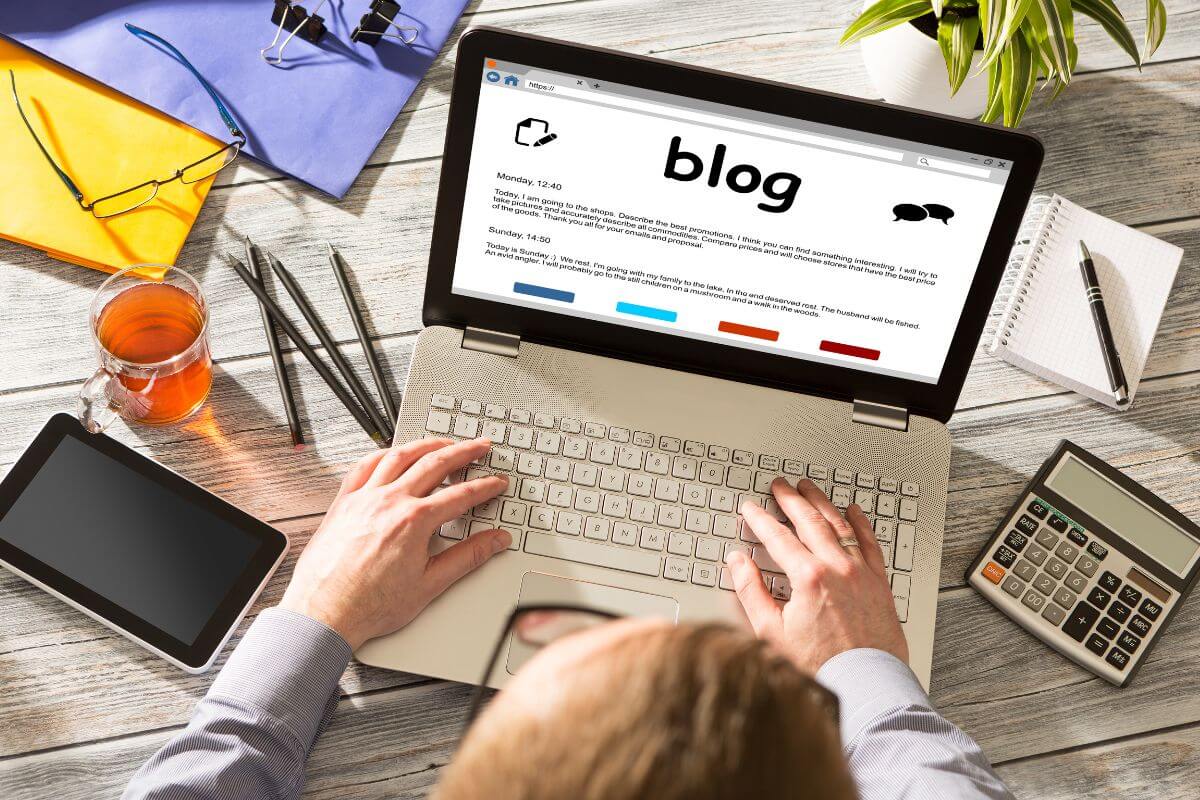 The image tells about papers, pens, a calculator, a tablet, a cup of tea, a plant, and a man's hand using a laptop with a screen display referring to starting a niche blog and monetizing it to make money from home.