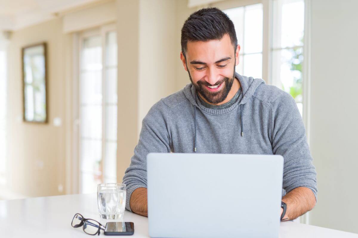 The photo shows a phone on the table, a glass of water, an eyeglass, and a man using a laptop which means to flip domain names for profit and to grow your income.