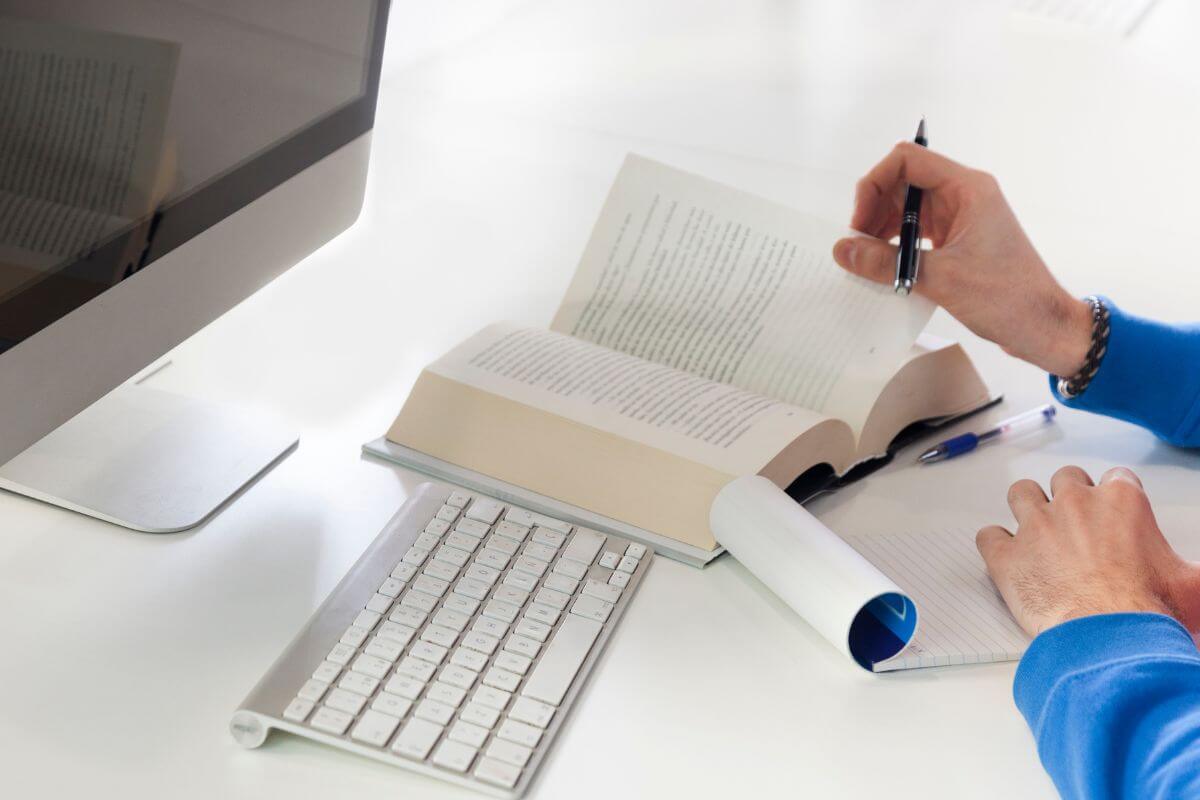 The photo shows a monitor, a keyboard, and a man's hand flipping the page of a book while writing in the notebook which refers to offering translation services that can bring in extra income.