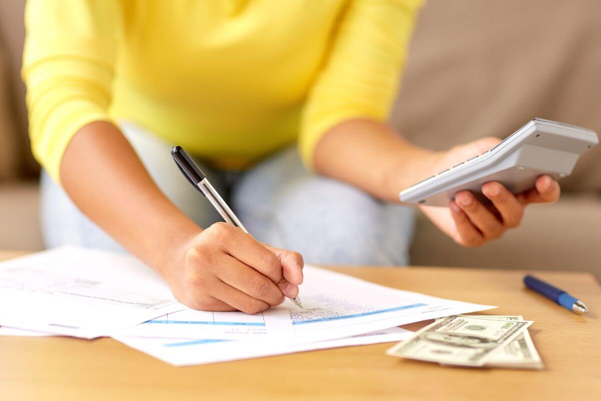 The picture shows dollar bills on the table, a pen, and a girl holding a calculator while writing on the papers referring to tracking all income and expenses helps you spot problem areas and see where your money goes.