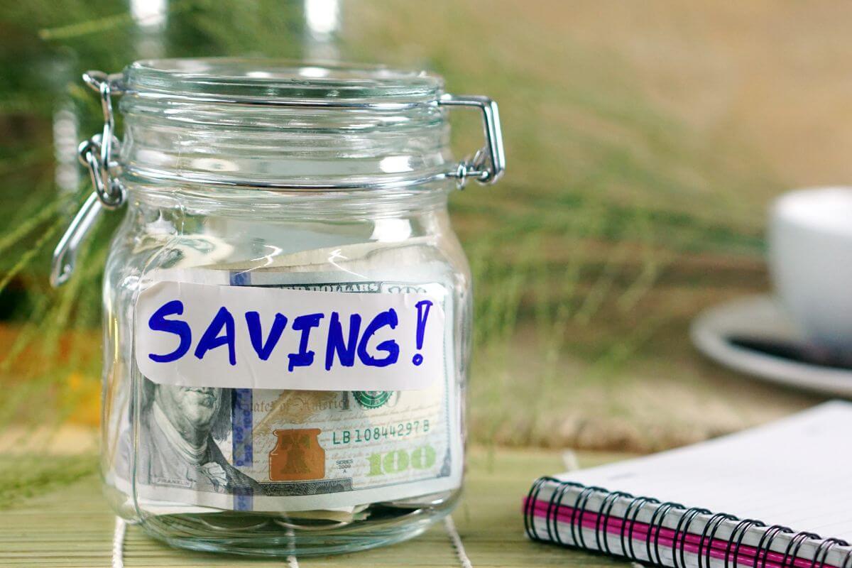 The image shows a notebook and a jar filled with coins and dollar bills with a note referring to automate savings and ensuring you're building wealth every month.