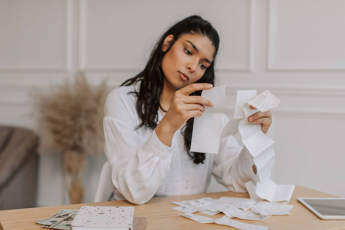 The picture shows dollar bills on the table, a notebook, a tablet, and a girl checking the receipts which refers to track spending and helps you adjust your spending and stay on track with saving for the future.